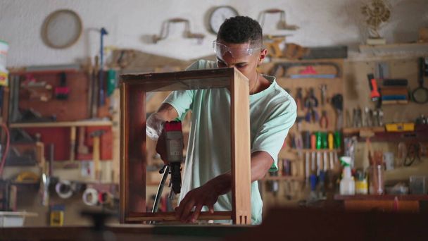 Participó en la escena artesanal de un joven carpintero brasileño negro que construye muebles en el taller, aprendiz con equipo de perforación, estudiante de carpintería de madera - Foto, Imagen