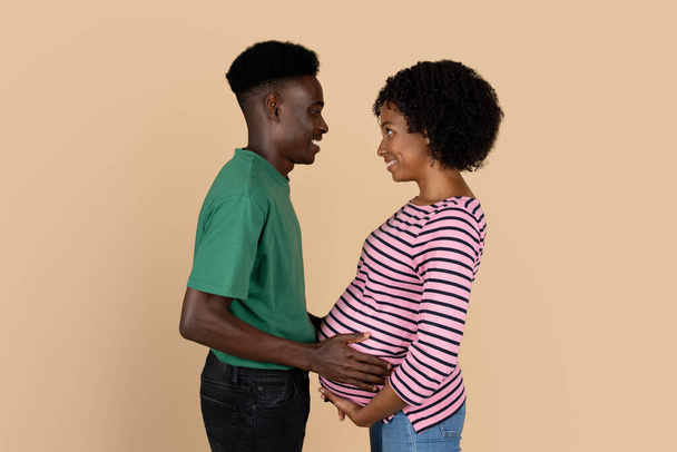Vrolijke duizendjarige zwarte man aanraken vrouw dikke buik, genieten van zwangerschap, het gevoel beweging, geïsoleerd op beige achtergrond, studio. Toekomstige familie, levensstijl en verwachte baby, advertentie, aanbod - Foto, afbeelding