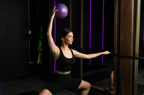 Ενεργό τακτοποίηση νεαρή γυναίκα άσκηση με μια μπάλα γυμναστικής, ενώ η πρακτική κατά τη διάρκεια ενός μαθήματος barre κατάρτισης σε ένα υψηλό τέλος γυμναστήριο - Φωτογραφία, εικόνα