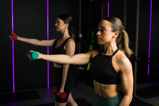 Atractivas mujeres de fitness levantando pesas de mancuerna y ejercitándose juntas durante una clase de entrenamiento funcional - Foto, imagen
