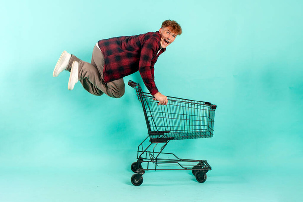 Genç çılgın adam alışveriş arabasıyla mavi izole edilmiş arka planda süpermarketten atlıyor ve seviniyor. Erkek müşteri hızla alışverişe koşuyor. - Fotoğraf, Görsel