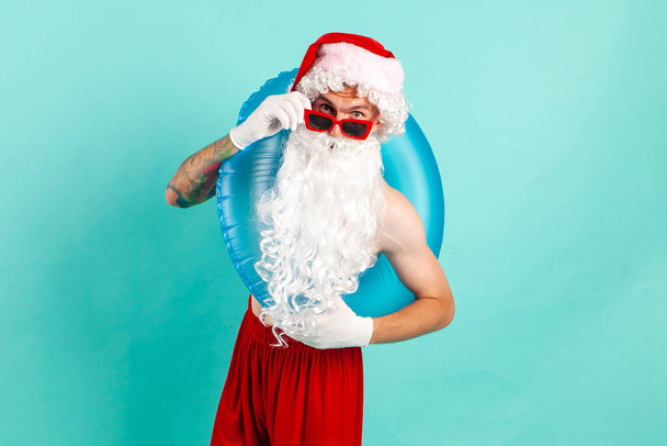 Санта-Клаус в костюме с надувным купальным кольцом в солнцезащитных очках лежит на пляже на синем изолированном фоне, Санта-турист отправляется в отпуск на море - Фото, изображение