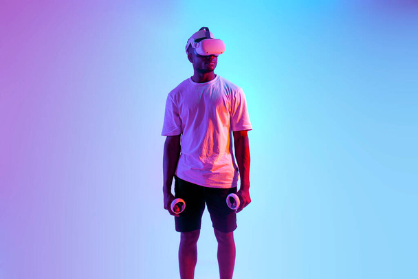 Αφροαμερικάνος στέκεται σε γυαλιά εικονικής πραγματικότητας σε νέον φωτισμό, σοβαρός τύπος σε vr γυαλιά κατέχει joysticks, αντιγραφή χώρου - Φωτογραφία, εικόνα