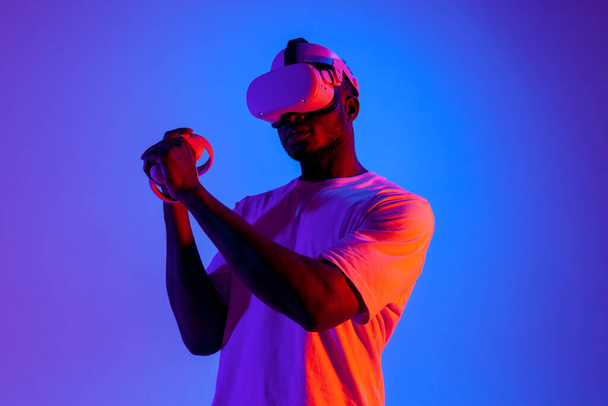 hombre afroamericano en gafas de realidad virtual apuntando y disparando en iluminación de neón, chico gamer jugando y luchando en gafas vr con joysticks - Foto, imagen