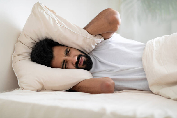 Não consigo dormir. Retrato de homem indiano jovem irritado deitado na cama e cobrindo os ouvidos com travesseiro, audição e sofrimento de som muito alto, cara oriental millennial irritado cansado de vizinhos barulhentos - Foto, Imagem
