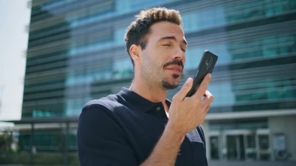 Serious man talking dynamic of mobile phone at glass building place closeup. Handsome latin business person dictating audio message using smartphone. Confident boss creating voice sms at downtown - Footage, Video