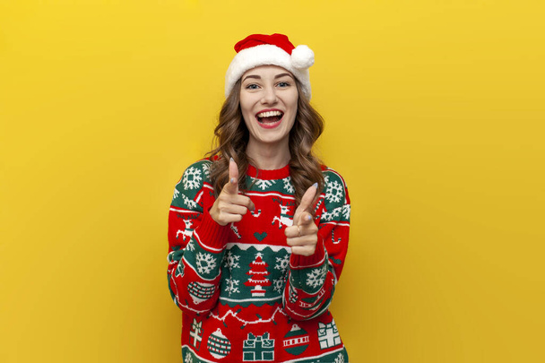 Je te choisis. jeune fille en pull de Noël et santa claus chapeau sourit et pointe vers la caméra sur fond jaune isolé, femme en vêtements de Noël pointe vers l'avant avec ses mains - Photo, image
