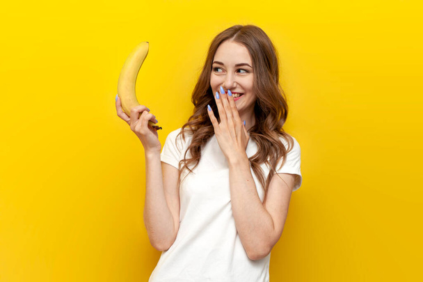 niña sosteniendo plátano pequeño y burlándose de fondo aislado amarillo, mujer presenta concepto erótico e íntimo, concepto de tamaño sexy - Foto, imagen