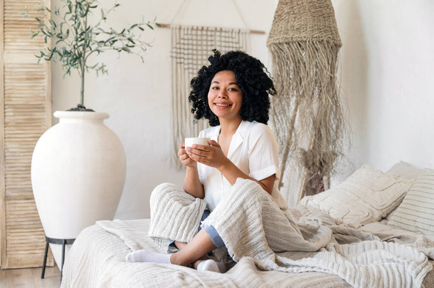 Şık ev kıyafetleri içinde gülümseyen Afro-Amerikan kadın keten tekstil ile rahat bir yatakta oturuyor ve elinde bir fincan sabah kahvesi tutuyordu. Bohem dekoratif elementlerle dolu rahat bir yatak odası.. - Fotoğraf, Görsel
