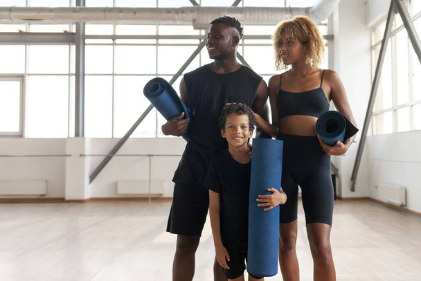 Spor kıyafetli Afro-Amerikan ailesi yoga minderleriyle spor salonunda duruyor ve gülümsüyor, anne baba ve oğul spor yapıyor. - Fotoğraf, Görsel