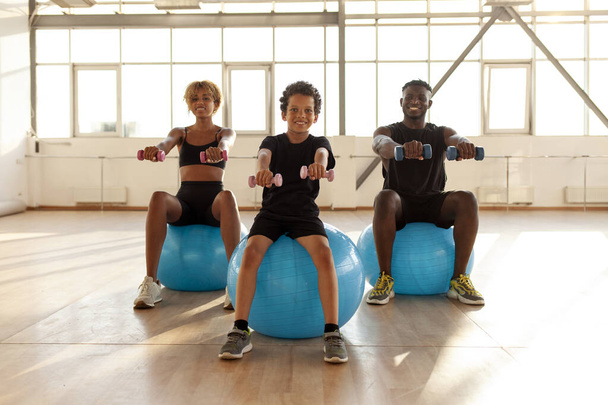 sporty african american family is doing pilates on fitness ball in the gym, boy with his parents is holding dumbbells and doing exercises, mom dad and son are warming up - Photo, Image