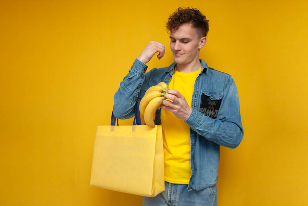 młody facet kupujący z tkaniny eko torba kupuje banany i umieszcza je w wielokrotnego użytku nieplastikowej torbie na żółtym tle, ekologia koncepcja - Zdjęcie, obraz