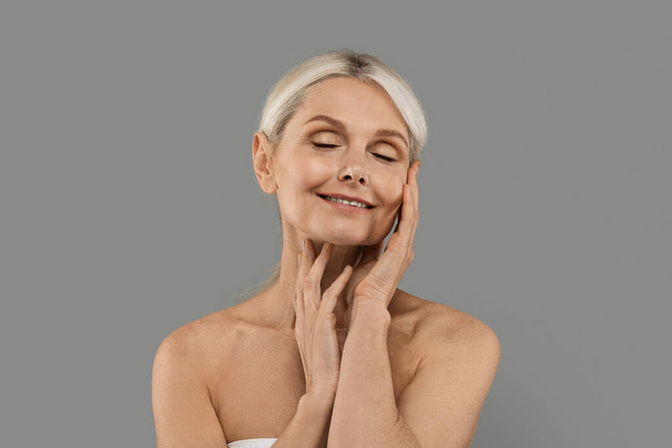 Kosmetyki przeciw starzeniu się. Senior Woman With Beautiful Soft Skin pozowanie na szarym tle, Atrakcyjne Dojrzałe Kobieta dotykając jej skóry i uśmiech, Korzystanie z zabiegów pielęgnacji skóry, Kopiuj przestrzeń - Zdjęcie, obraz