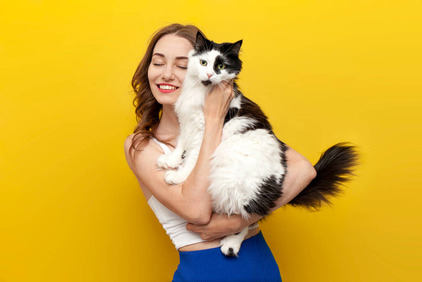 黄色で隔離された背景と笑顔に黒と白の猫を保持する若いかわいい少女, ペットが着色された背景にポーズする女性, 動物の世話と愛 - 写真・画像