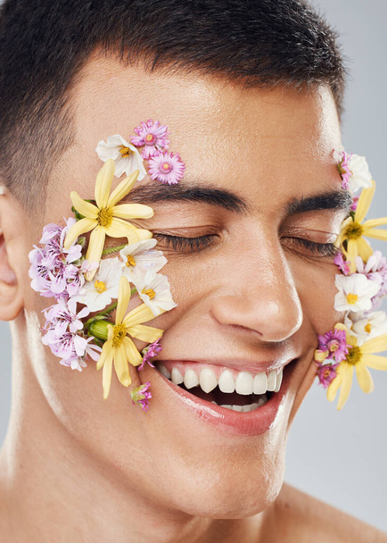 Smile, beauty and a man with flowers on a studio background for creative aesthetic. Happy, plant and a male model with floral art for facial wellness and dermatology isolated on a studio backdrop. - Fotografie, Obrázek