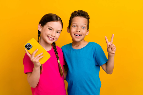 Photo of excited pretty preteen schoolkids dressed t-shirts texting modern gadget showing v-sign isolated yellow color background. - Foto, Imagen