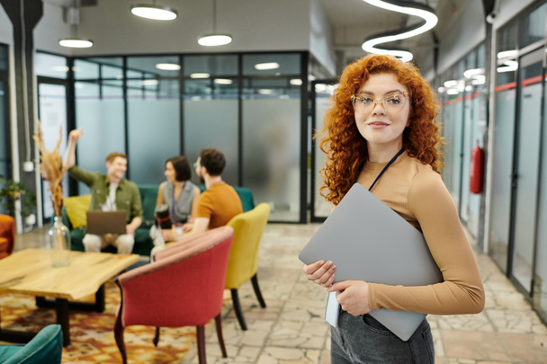 redhead team lead with laptop near colleagues talking on blurred background, headshot - Photo, Image