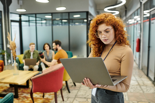 vörös hajú üzletasszony nézi laptop, míg a csapat beszél homályos háttér a modern irodában - Fotó, kép