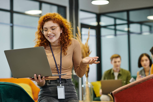 successful redhead businesswoman with laptop near blurred colleague in modern office space - Фото, зображення