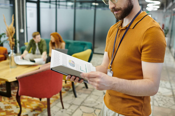 бородатый бизнесмен смотрит на графики, менеджеры разговаривают на размытом фоне в современном офисе - Фото, изображение