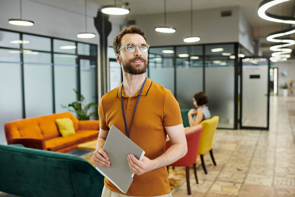 Lächelnder bärtiger Geschäftsmann mit Brille, Dokumente in der Nähe verschwommener Kollegen im Coworking-Büro - Foto, Bild