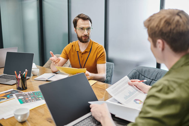 bearded entrepreneur talking to colleague with documents near laptops and graphs at workplace - 写真・画像
