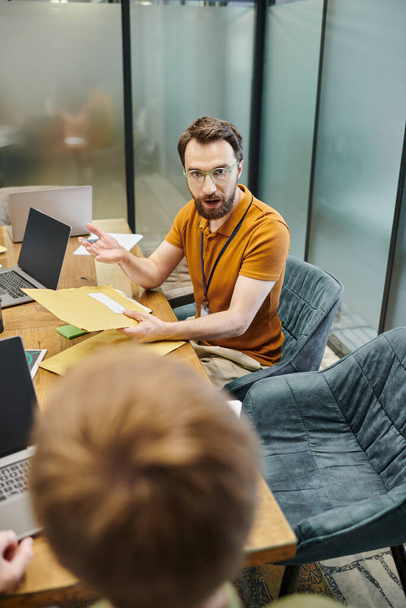 entrepreneur barbu avec des enveloppes parler à des partenaires d'affaires flous, ordinateurs portables, lieu de travail moderne - Photo, image