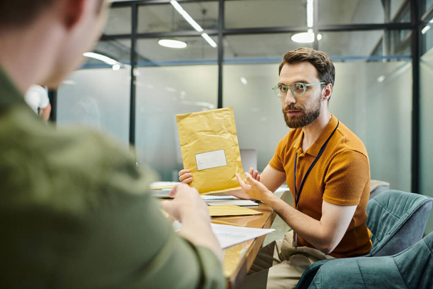 бородатий бізнесмен показує конверт розмитому колезі, сучасному офісу, робочому місці - Фото, зображення