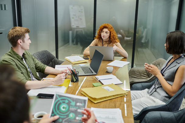 рыжая деловая женщина разговаривает с коллегами возле документов и ноутбуков в офисе, обсуждая стартап - Фото, изображение