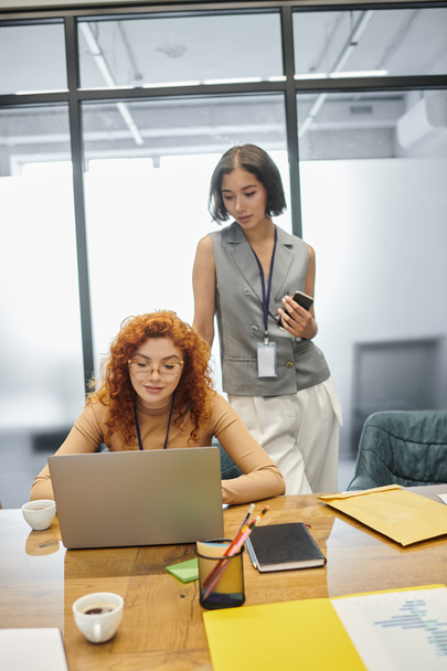 jovem liderança da equipe com smartphone perto de gerente ruiva trabalhando no laptop no escritório moderno - Foto, Imagem