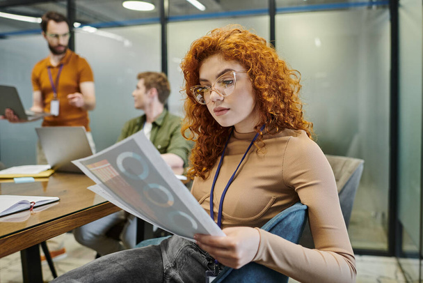 Przemyślana rudowłosa bizneswoman z infografiką, zamazani koledzy rozmawiający w nowoczesnym biurze - Zdjęcie, obraz
