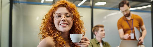 Gözlüklü kızıl saçlı girişimcinin portresi, bulanık meslektaşlarının yanında kahve fincanı, pankart. - Fotoğraf, Görsel