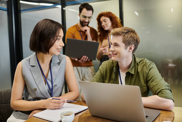 glimlachende managers praten in de buurt van laptop en collega 's op wazige achtergrond, zakelijke samenwerking - Foto, afbeelding