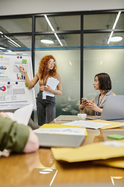 рыжая деловая женщина, указывающая на инфографику на флип-чарте рядом с коллегами в конференц-зале - Фото, изображение