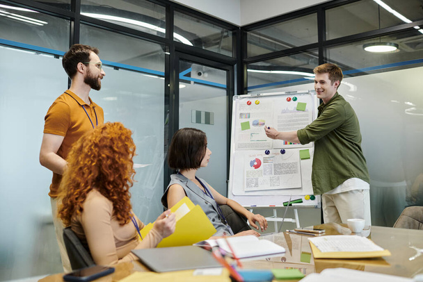 Geschäftsprojekt planen, lächelnder Unternehmer zeigt auf Grafiken in der Nähe von Kollegen im Besprechungsraum - Foto, Bild