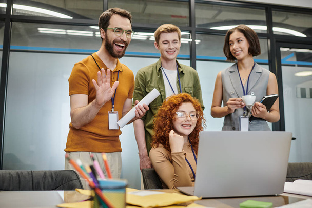vrolijk bebaarde ondernemer zwaaien hand tijdens video conferentie in de buurt van zakelijke partners in kantoor - Foto, afbeelding