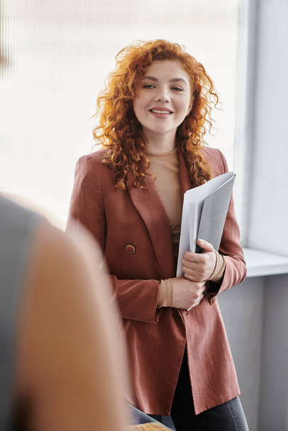 веселая рыжая деловая женщина с документами в офисе, профессиональный портрет - Фото, изображение