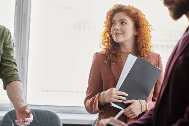 young redhead businesswoman with documents smiling near colleagues in contemporary office - Photo, Image