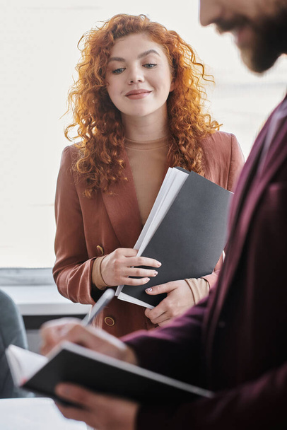 redhead businesswoman with documents near blurred colleague writing in notebook in modern office - Zdjęcie, obraz