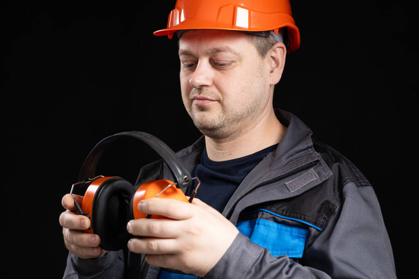 Builder in a protective helmet with noise-cancelling headphones in his hands on a black background. - Foto, afbeelding