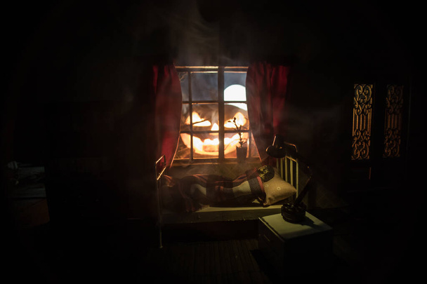Pomysł na Halloween. Realistyczna sypialnia domku dla lalek z meblami i oknem w nocy. Przerażony człowiek w łóżku z wielką świecącą dynią. Skupienie selektywne - Zdjęcie, obraz