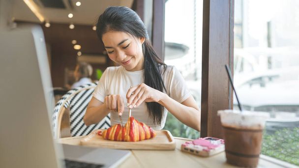 Az emberek pihennek a kávézóban. Boldog mosoly arca fiatal felnőtt ázsiai nő eszik croissant kenyeret. Az asztalon laptoppal. Bent ül az ablaknál napközben.. - Fotó, kép