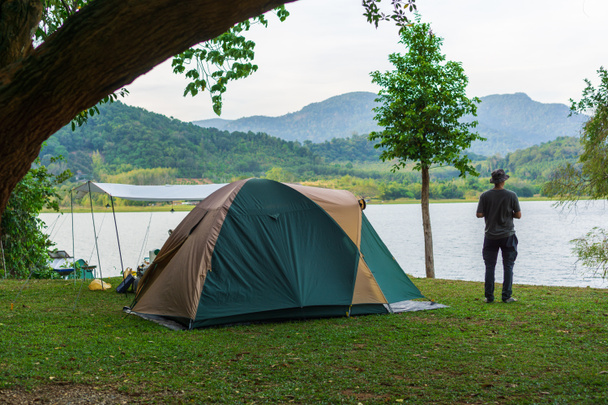 Азиатский мужской туристический кемпинг палатка на отдыхе на озере, лагерь на зеленой траве под большими деревьями и распространение брезента, красивое озеро и вид на горы утром, отдых. - Фото, изображение