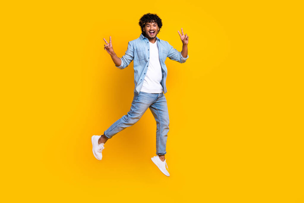 Full length photo of positive funny guy dressed denim jacket jumping high showing v-signs isolated yellow color background. - Zdjęcie, obraz