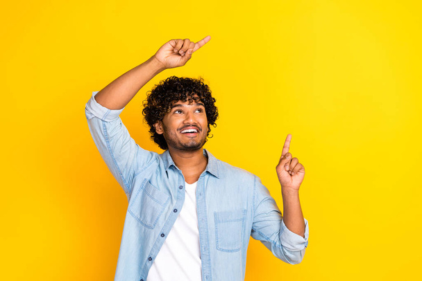 Photo of good mood man with curly hairdo dressed denim shirt directing look at sale empty space isolated on yellow color background. - Fotó, kép