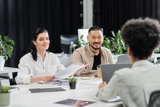 vrolijke interraciale zakenmensen kijken naar vrouwelijke collega tijdens het werken in moderne coworking - Foto, afbeelding