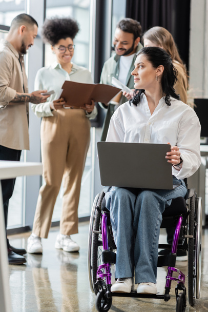 disabled businesswoman in wheelchair using laptop near blurred multiethnic team, inclusion concept - Fotoğraf, Görsel