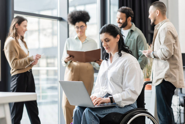 niepełnosprawna bizneswoman na wózku inwalidzkim przy użyciu laptopa w pobliżu rozmazanych współpracowników, włączenie - Zdjęcie, obraz