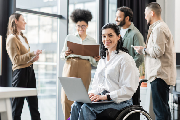 happy disabled businesswoman in wheelchair using laptop near blurred multiethnic colleagues - Foto, Bild