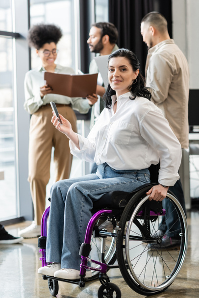 szczęśliwa niepełnosprawna bizneswoman na wózku inwalidzkim patrząc na kamerę w pobliżu zamazanych wielonarodowych kolegów - Zdjęcie, obraz